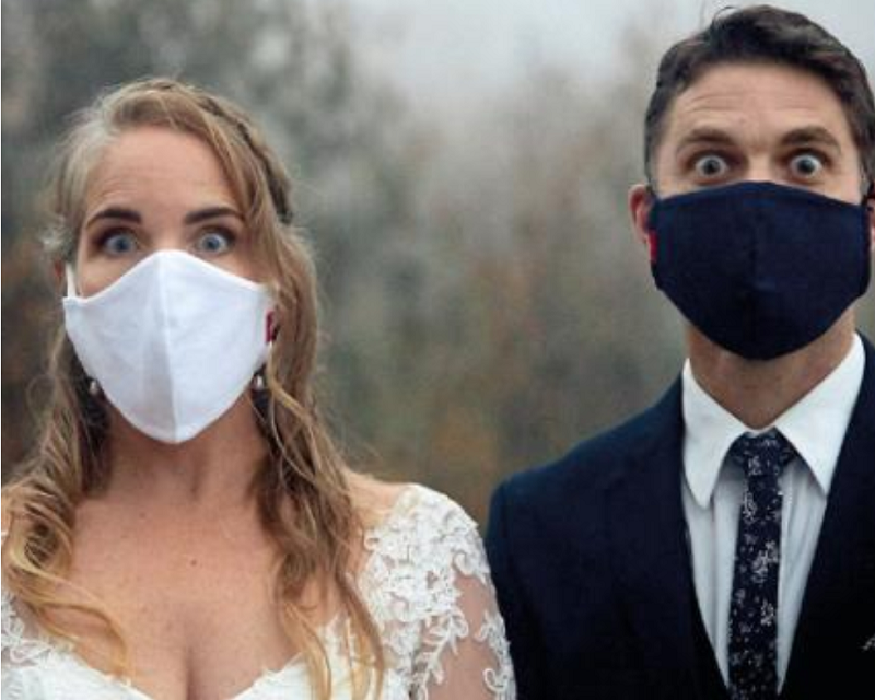 A bride and bridegroom stand next to each other facing the camera, both are wearing a mask covering their nose and mouth